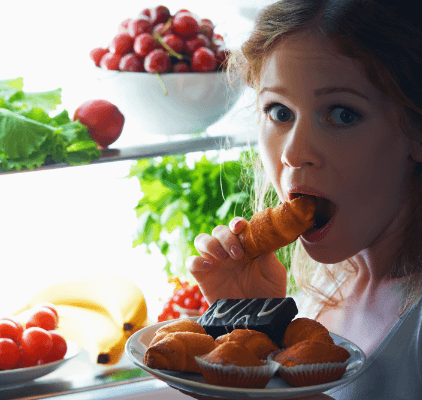 A woman eating a croissant in front of a fridge filled with veggies. 